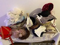 Vintage ladies hats and gloves displayed on a cloth-covered surface, showing variety of styles, colors, and materials including feathers, lace, netting, pearls, and sequins.