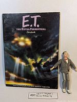 Photo showing E.T. Storybook cover with colorful lights and title, with Peewee Herman toy figure next to it.