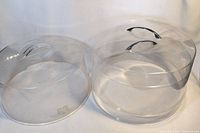Photo showing two clear plastic cake covers, one with handle and one without, both round and transparent, placed side by side.