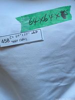 Close up photo of white fabric with a green tape note reading '64x64 x 5' and a label reading '458 5x 64" x 64" white table cloths'