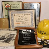Photo showing framed certificates, wooden plaque, metal plaque, vintage black and white photo, and yellow CNR hard hat.