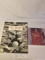 Two books side by side on a white cloth background: larger M.C. Escher The Graphic Work book and smaller Klimt art book.