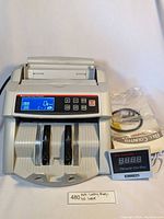 Front view of multi-currency bill counter with digital display screen, external digital display, power cord, cleaning brush, and user manual next to it on white background.