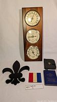 Photo showing wooden wall-mounted set of barometer, thermometer, and hygrometer, black fleur de lis metal decoration, small tricolor item, black leather pouch with fleur de lis emblem, and 1944 tide tables book with blue cover.