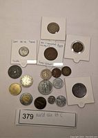 Overall layout of 18 world coins on flat white surface with two coins mounted in white cardboard holders with labels about rarity and origin