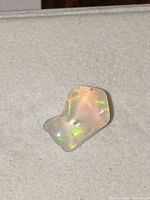 Close-up photo of a single small polished opalite gemstone showing glowing green and yellow iridescent flashes on a neutral light background.