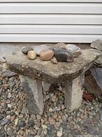 Three-legged natural limestone garden table with flat stone top and several natural stones placed on top, shown outdoors on gravel ground.