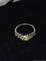 Front angled view of silver-colored rhodium plated ring with heart-shaped green peridot center and small green and white stones on sides.