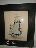 Framed print showing two girls in Dutch traditional clothing and wooden shoes, with hand-tinted or watercolor style color behind glass in an oak wood frame.