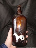 Bottle held in hand showing front with white horse silhouette decal on amber glass