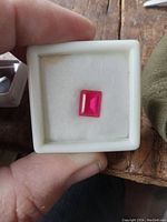 Lab grown ruby gemstone in a white display box, showing shape, cut and color with corner chip.