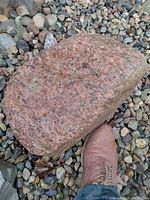 Pink granite landscaping boulder placed on gravel, showing rough textured surface and moderate size compared to a boot for scale.