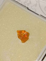 Image showing a polished, irregular-shaped dark orange fire opal with visible rainbow flecks inside, placed on a pale textured background.