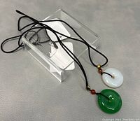 Two round jade pendants on black cords laid on grey textured surface, showing full pendants and cords with bead accents.