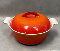 Front view of medium size Belgium cast iron pot with orange to red enamel finish and white handles, lid on.