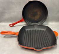 Photo of square grill pan and round skillet stacked showing cast iron surfaces and enamel-coated orange handles.