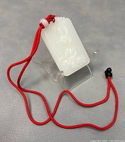 Front view of rectangular white jade pendant carved with Guanyin, attached to red cord with beads, placed on a clear stand.