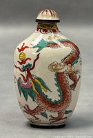 Front view of the small oval white enamel snuff bottle painted with a red and green dragon chasing a flaming pearl, showing the intricate hand-painted details and the removable lid with matching design.