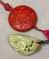 Close-up view of the red cinnabar pendant with detailed floral carving and the carved jade pendant, highlighting the craftsmanship and cords.