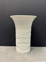 Front view of the white porcelain vase showing embossed wave and leaf pattern wrapping around body with flared top opening.