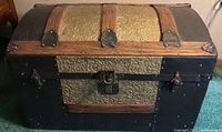 Front view of antique dome-top trunk showing embossed gold floral metal panels with wood and black metal reinforcements and central lock clasp.