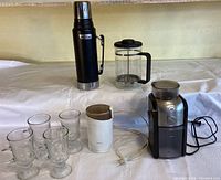 Lot contents showing all items: Stanley thermos, French press, electric coffee grinder, milk frother, and four glass mugs on a white draped surface