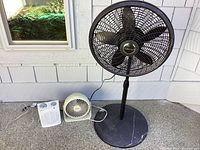 Tall Lansko fan, small Honeywell fan, and Holmes heater on patio floor in front of siding wall and window.