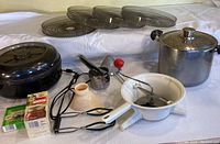 Photo showing electric food dehydrator, pressure cooker, tongs, hand crank sieve, food press, and seasoning packets on white cloth