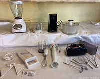Full view of handheld kitchen appliances including blender, immersion blender, food chopper, hand mixer with attachments, can opener and salad spinner laid out on a white cloth