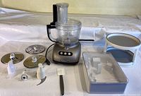 Full view of KitchenAid food processor with multiple slicing/shredding discs, various blades, spatula, storage container, and stand.
