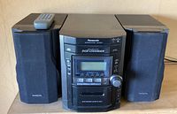 Front view of the Panasonic stereo system with 5-disc CD changer, cassette deck, and remote control on top, flanked by two black Panasonic speakers.