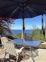Full setup of patio table with glass top, three chairs around, blue umbrella opened and inserted in table hole, outdoor deck setting with ocean view