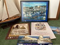 Photo showing framed signed poster, two unframed posters, calendar, puzzle box, and wooden boat replica arranged on the floor near wall