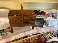 Wooden cantilever sewing box closed with rolls of upholstery fabric and sewing tools around it.