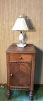 Full view of vintage wood nightstand with a lamp on top. Nightstand has a cabinet door and a drawer with a round knob. Lamp is white with brass and milk glass elements, topped with a white shade.