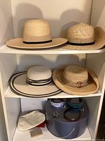 Four straw hats arranged on two shelves: two hats on top, two on middle shelf. Bottom shelf has a small white beaded clutch, a silver metal card case, two round boxes, three headbands, and an enameled flower pin.