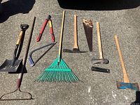 Full view of yard and garden tools arranged on concrete surface including shovel, rake, loppers, hand saw, axe, pick mattock, pruning saw, and hoe.