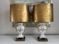 Two matching porcelain lamps with gold foil shades on a surface, framed by artwork in background