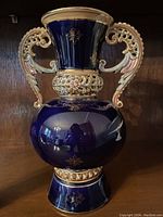 Front view of the cobalt blue vase with gold gilt highlights and decorative handles on wooden shelf.