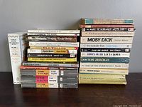 Photo showing a large stack of classic paperback books featuring titles by Hemingway, Steinbeck, Brontë, Melville, Hardy, Fitzgerald, and others, arranged with visible wear and tear.