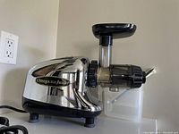 Side angle view of the Omega juicer showing chrome body and black base feet, with the funnel and container attached.