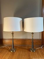 Pair of UpCountry brass and black lamps with white drum shades placed on wooden floor against wall.