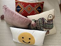Four decorative pillows arranged on a light striped fabric surface showing variety of patterns and colors including geometric, embroidered bird, smiley face, and solid mauve with tassels.