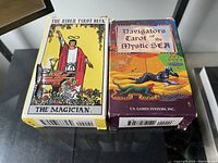 Two boxes of tarot cards side by side on glass table. Left box is The Rider Tarot Deck with The Magician on front. Right box is Navigators Tarot of the Mystic Sea with underwater themed artwork.