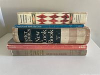 Stack of six vintage books showing the Mennonite Community Cookbook on top and Better Homes and Gardens New Cook Book thicker volume in middle