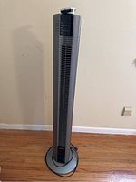 Full view of silver and black Lasko tower fan on wooden floor near wall.