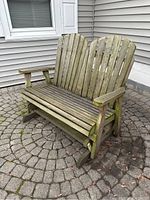 Front-side view of wooden Adirondack glider bench on stone patio with weathered wood condition and some moss growth.
