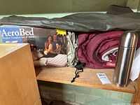 Queen Aero Bed air mattress box, carrying pouch, Coleman sleeping bag, and stainless flask on a wooden shelf.