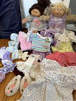 Two vintage Cabbage Patch dolls sitting with assorted children's clothing and shoes arranged in front of them on the floor.