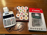 Canon P-170-DH printing calculator with a roll of paper tape installed, a second calculator still in packaging, and multiple rolls of paper tape refills on wooden surface.